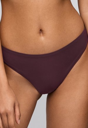 Middenstuk van een vrouw die effen donkerpaarse bikinibroekjes draagt met zichtbare navel en linkerhand ontspannen langs haar zijde.