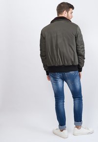 Grüne Bomberjacke mit einem Fellkragen, ausgestattet mit einer gesteppten Textur, gerippten Bündchen und Saum. Tragen mit blauen Jeans und weißen Turnschuhen.