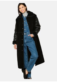 Zwarte schapenvachtjas met leren mouwen, oversized ontwerp, knoopsluiting, gedragen over een denim jumpsuit, met zakken en een hoge kraag.