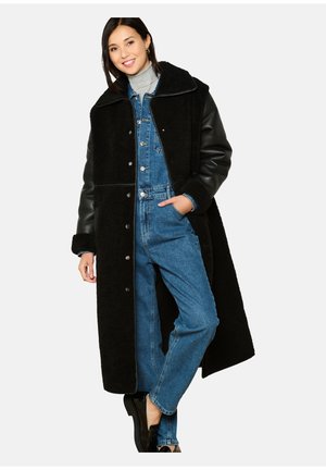 Zwarte schapenvachtjas met leren mouwen, oversized ontwerp, knoopsluiting, gedragen over een denim jumpsuit, met zakken en een hoge kraag.