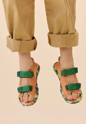 Sandalen mit pfirsichfarbenem Obermaterial, grünen Klettverschlüssen und mehrfarbigem Muster an der Sohle. Weiches Material, offenes Design und sichtbare Zehen.