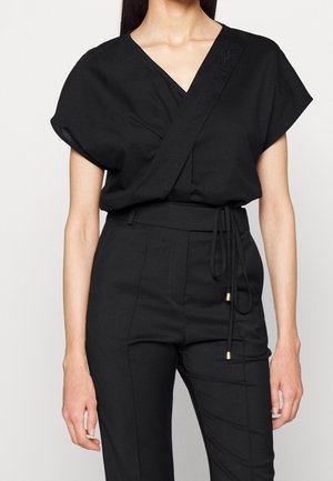 Woman wearing a black short-sleeve wrap-style top tucked into high-waisted black pants with a tied belt.