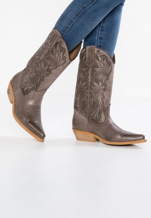 Nogi w brązowych skórzanych kowbojskich butach z kwiatową haftowaną ozdobą i drewnianymi obcasami, zestawione z niebieskimi dżinsami, na białym tle.