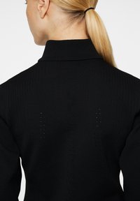 Svart stickad tröja med hög krage, texturerat tyg och subtila perforeringar. Textdetalj synlig längs ryggen.
