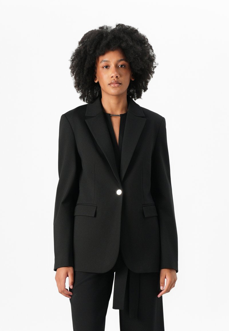 Zwarte blazer met een enkele knoopsluiting, notch-revers en twee voorzakken. Glad, gestructureerd stof; gecombineerd met zwarte broek.