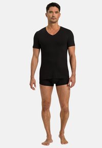 Černé tričko s krátkým rukávem a výstřihem do V z hladkého materiálu, spárované s černými přiléhavými boxerkami. Model stojí bosý proti jednoduchému pozadí.