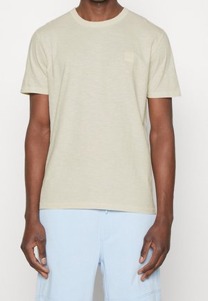 Mężczyzna ubrany w beżowy t-shirt z krótkim rękawem z małym logo na piersi oraz jasnoniebieskie casualowe spodenki, stojący na białym tle.