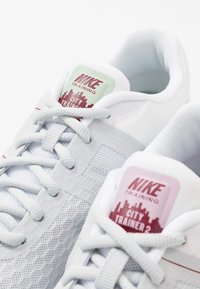 Hvide mesh træningssko med lysegrå detaljer. Funktionen inkluderer strukturerede sider og farverige hælklapper med branding. Snørebåndene er lysegrå.