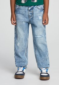 Jeans in denim azzurro chiaro con un fit rilassato, caratterizzati da dettagli di usura e un classico design a cinque tasche. Indossati con sneaker blu e bianche.