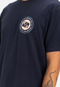Camiseta de algodón azul marino con un logo redondo que presenta una 'S' estilizada en el centro y texto alrededor. Mangas cortas, diseño de cuello redondo.