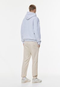 Felpa grigia con cappuccio, pantaloni beige e sneakers chiare. Vestibilità ampia, tessuto testurizzato, con dettagli di cucitura visibili sui pantaloni.