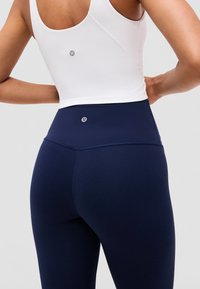 Person trägt ein weißes ärmelloses Top und marineblaue Leggings mit hoher Taille und Logos auf dem Rücken, die Hände auf den Hüften ruhend.