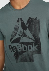 Zelené tričko s velkým černým logem Reebok a grafickým designem na hrudi, nošené osobou oříznutou u krku.
