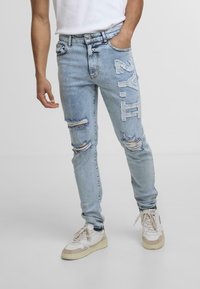 Hele sinine teksapüksid, millel on kulumisjäljed ja külgel trükk. Sobiv disain kerge elastusega. Kaasa kantud heledate sneakeritega.