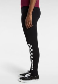 Person som bär svarta Vans leggings med vit rutmönstring och logotyp längs vaden, i kombination med svarta och vita Vans sneakers.