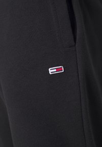 Černé tepláky vyrobené z bavlněné směsi, s malým vyšitým logem v červené, bílé a modré barvě na levé straně.