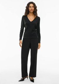 Svart långärmad topp med V-ringning, gjord av ett glittrande texturerat material, kombinerat med högmidjade svarta byxor och eleganta, glänsande skor.