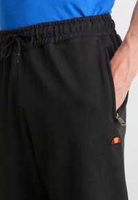 Pantalones deportivos negros con cinturilla elástica, con cordón y un bolsillo con cremallera. Incluye un pequeño acento de logo colorido.