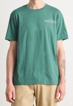 T-shirt vert à manches courtes avec petit logo blanc GANT 1949 sur la poitrine gauche, porté par une personne avec un avant-bras droit tatoué et un pantalon beige.
