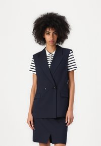 Marineblå ærmeløs blazer med dobbelte knapper og sidelommer, lagt over en svart og hvit stripete skjorte, paret med et matchende skjørt.