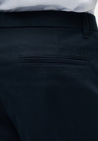 Prim-plan cu pantaloni negri croiți, evidențiind un singur buzunar spate cu fanta, cu un logo subtil Calvin Klein embosat deasupra acestuia.