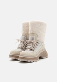 Bottines beige à cheville avec revers en fausse fourrure, accents en maille argentée, semelle plateforme robuste et fermeture à lacets. En suède texturé et matériaux lisses.