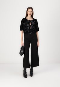 Crni top sa sequinsom i kratkim puhovim rukavima, s mašnom za zatvaranje sprijeda, uparen s crnim hlačama širokog kroja i čizmama s petom.