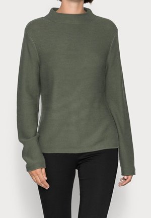 Femme portant un pull vert olive texturé à manches longues avec un col montant, associé à un pantalon noir, sur un fond clair uni.