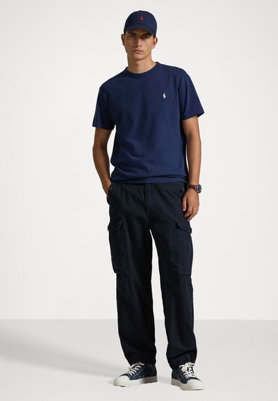 Marineblå bomull crew-neck t-skjorte med liten rød logo, kombinert med mørke cargo-bukser som har sidelommer og marineblå kanvas-sneakers.