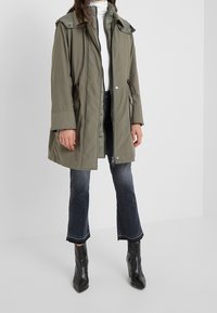 Olivgrön parkas med dragkedja framtill, fickor med knappar, justerbar snörning i midjan och avtagbar huva; kombinerad med mörka utsvängda jeans.