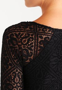 Schwarze Spitze mit komplexen Blumenmustern und geometrischen Formen. Sie hat eine durchsichtige Textur und einen runden Ausschnitt.