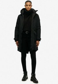 Czarna puchowa kurtka z wysokim kołnierzem, długimi rękawami i bocznymi kieszeniami, noszona na czarnym swetrze, w zestawieniu z czarnymi obcisłymi dżinsami i botkami.