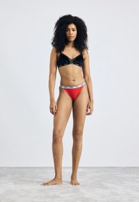 Black velvet bralette with a V-shaped design and logo band; red thong with a white logo waistband. Model stands on a light textured surface.