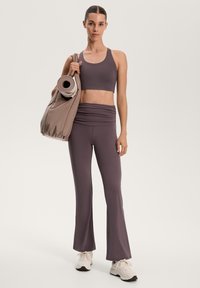 Könnyű, lila sportos szett, amely egy testhezálló crop topból és bővülő szárú leggingsből áll; bézs táskával és jógaszőnyeggel párosítva.