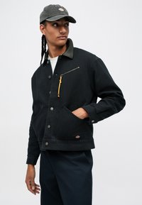 Νεαρός άνδρας με πλεξούδες, φορώντας μαύρο μπουφάν με τσέπη φερμουάρ και μαύρο καπέλο, κοιτάζοντας προς τα πλάγια, με το χέρι στην τσέπη, μπροστά σε λευκό φόντο.