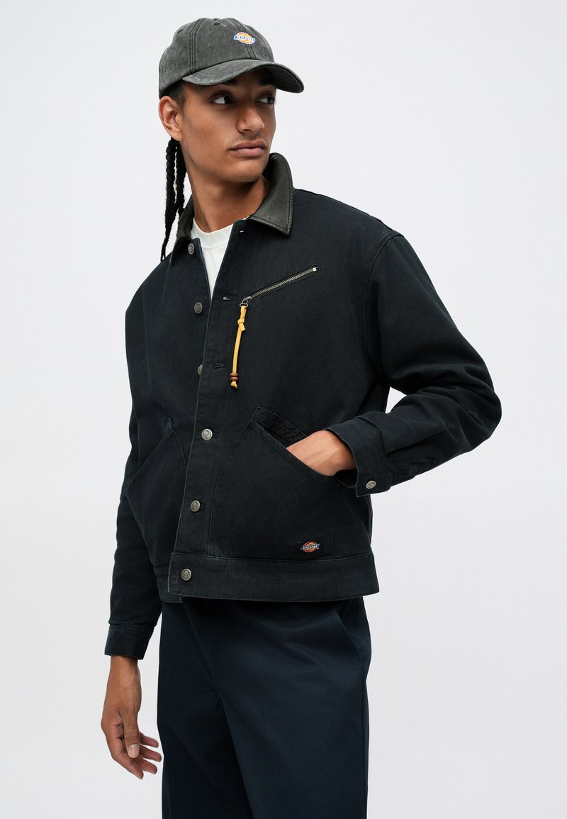 Young man with braided hair wearing a black jacket with zipper pocket and black cap, looking sideways, hand in pocket against white background.