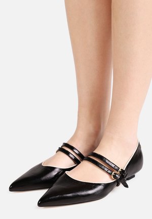 Stopy w czarnych balerinach z ostrym noskiem, podwójnymi cienkimi paskami z klamrami i niskim obcasem na jasnym tle.