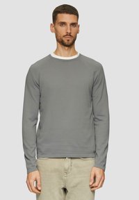 Maglietta grigia a maniche lunghe con scollatura a girocollo, caratterizzata da una texture a coste e un dettaglio del colletto interno bianco. Il modello indossa jeans chiari.