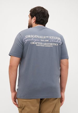 Siva bombažna majica z belim napisom na hrbtu: "ORIGINALS™ STUDIO Kopenhagen EST.1990 USTVARJENE ESTETIKE DNEVNI OSNOVNI OBLAČILA."
