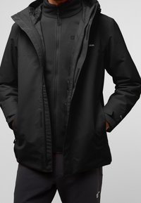 Hombre vestido con chaquetas negras de exterior en capas, con las manos en los bolsillos, mostrando cremalleras y detalles de la tela sobre un fondo liso.