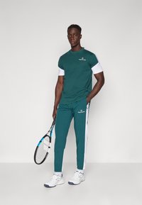 Grøn sportsbeklædning bestående af en T-shirt med korte ærmer og hvide detaljer samt matchende bukser med hvide sidebjælker. Tennisracket inkluderet.