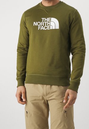 Mann trägt olivgrünes The North Face Sweatshirt und beige Cargohosen mit Reißverschlusstaschen vor schlichtem Hintergrund.