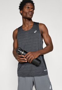 Μαύρο αθλητικό tank top με υφή, διαθέτει ανακλαστικό λογότυπο και πλαϊνές διχτυωτές επιφάνειες. Κρατώντας μια μαύρη φιάλη νερού. Γκρίζα σορτς.