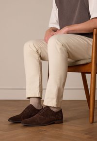 Brown suede dress shoes with flat laces, beige cotton trousers, and light grey socks, seated on a wooden chair against a neutral background.