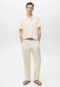 Maglietta polo a maniche corte di colore chiaro, pantaloni beige su misura, cintura marrone e sandali. Tessuto liscio, design semplice, vestibilità casual.