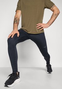 Black athletic leggings with a smooth texture, fitted design, and reflective logo detail, paired with an olive-green t-shirt and black running shoes.