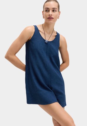 Mujer con vestido sin mangas azul oscuro y collar con colgante de piedra, de pie con las manos detrás de la espalda, mirando hacia un lado.