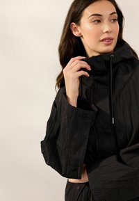 Veste noire texturée avec un col haut, une capuche à cordons de serrage et des manches surdimensionnées. Caractéristiques d'une fermeture à pression à l'avant et d'un éclat subtil.