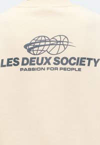 Μπεζ ύφασμα με μαύρο κείμενο "LES DEUX SOCIETY" και "PASSION FOR PEOPLE" κάτω από δύο αλληλοεπιδρώντα εικονίδια παγκόσμιου χάρτη και καλαθοσφαίρισης.
