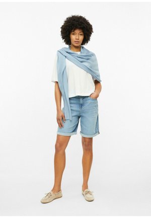 Femme debout avec une main dans la poche, portant une chemise blanche, un short en jean, un pull bleu sur les épaules et des mocassins beiges.
